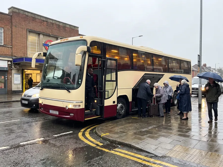 A cream and burgundy touring coach at the kerb in steady rain on the road outside Hornchurch Underground station, an older tour group boarding under umbrellas while the driver loads a case into the luggage bay